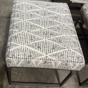 Gray and ivory patterned upholstered ottoman with black metal base and cushioned top.