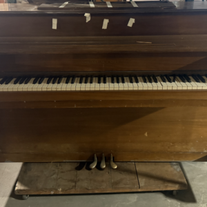 Vintage upright piano in warm wood finish with full keyboard and three pedals.